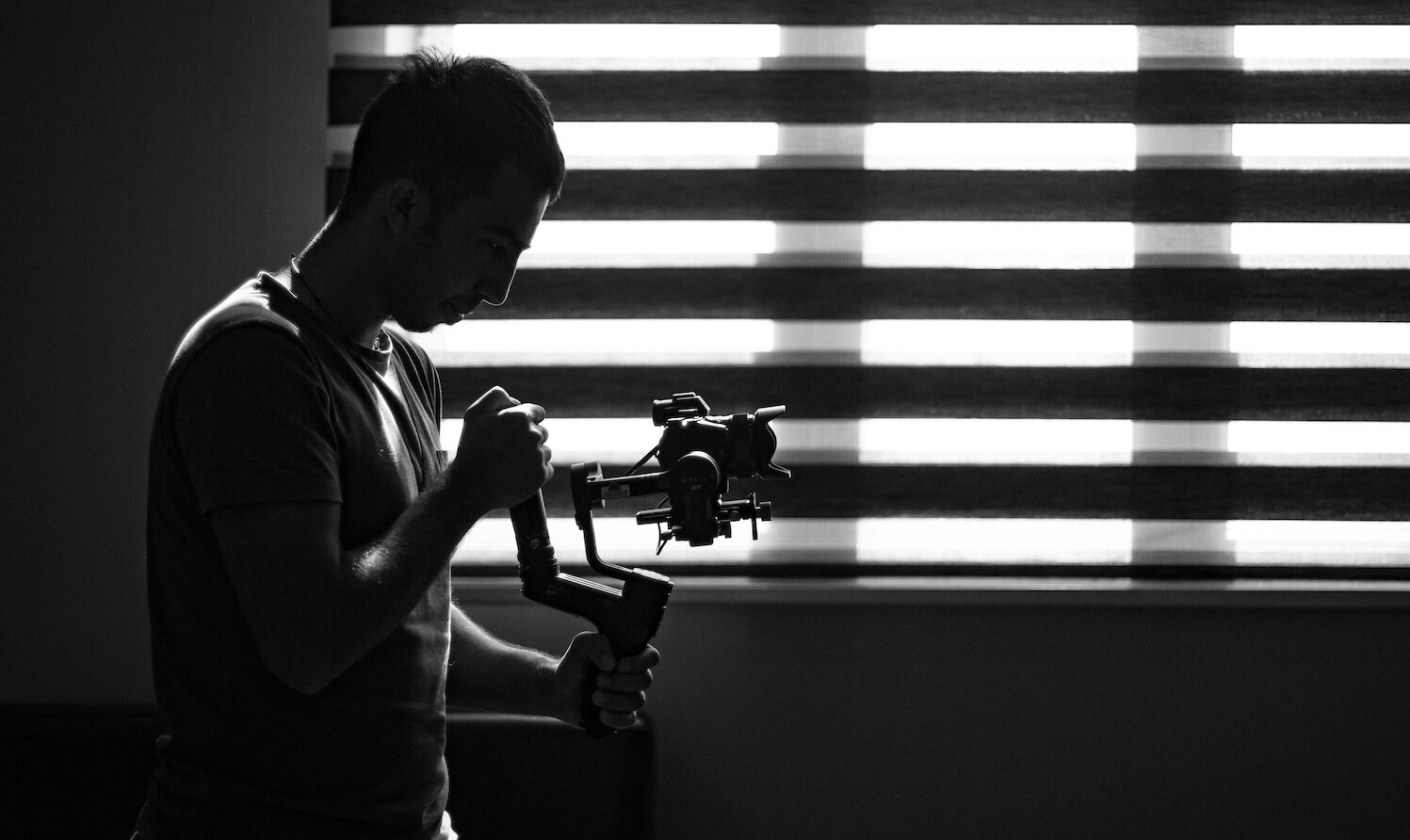black and white image of man standing sideways holding camera in front of blinds
